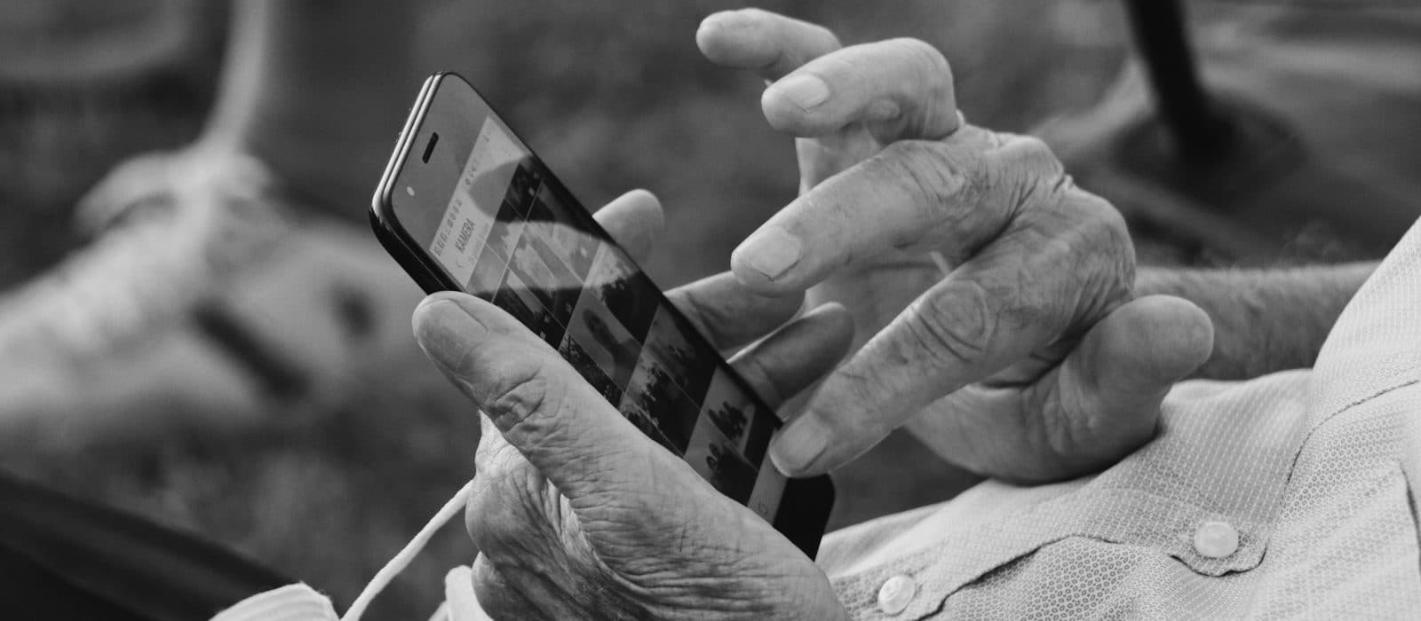A black-and-white close-up of an older person's hands holding a smartphone and reaching to tap the screen.