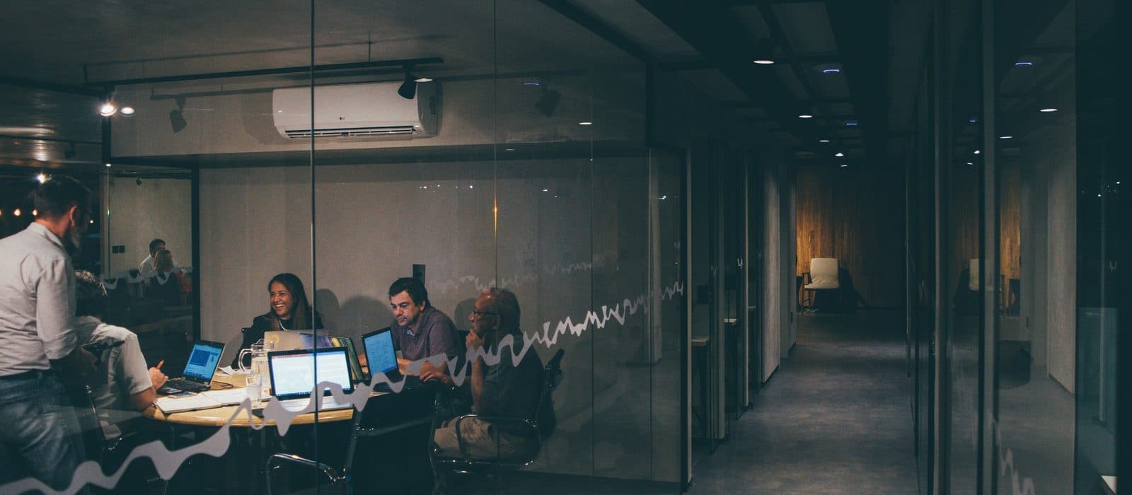 A small team working late around a table of open laptops, seen at night through the glass wall of an office.