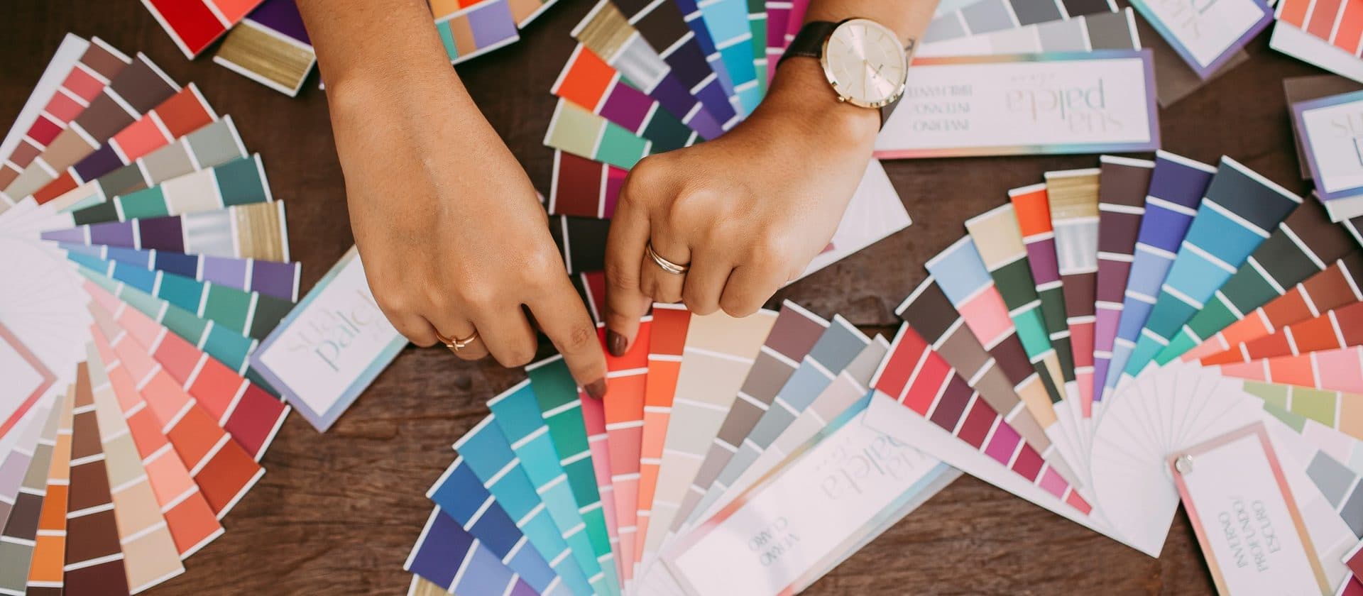 Hands choosing from colour swatch fans and sample cards spread across a wooden desk.