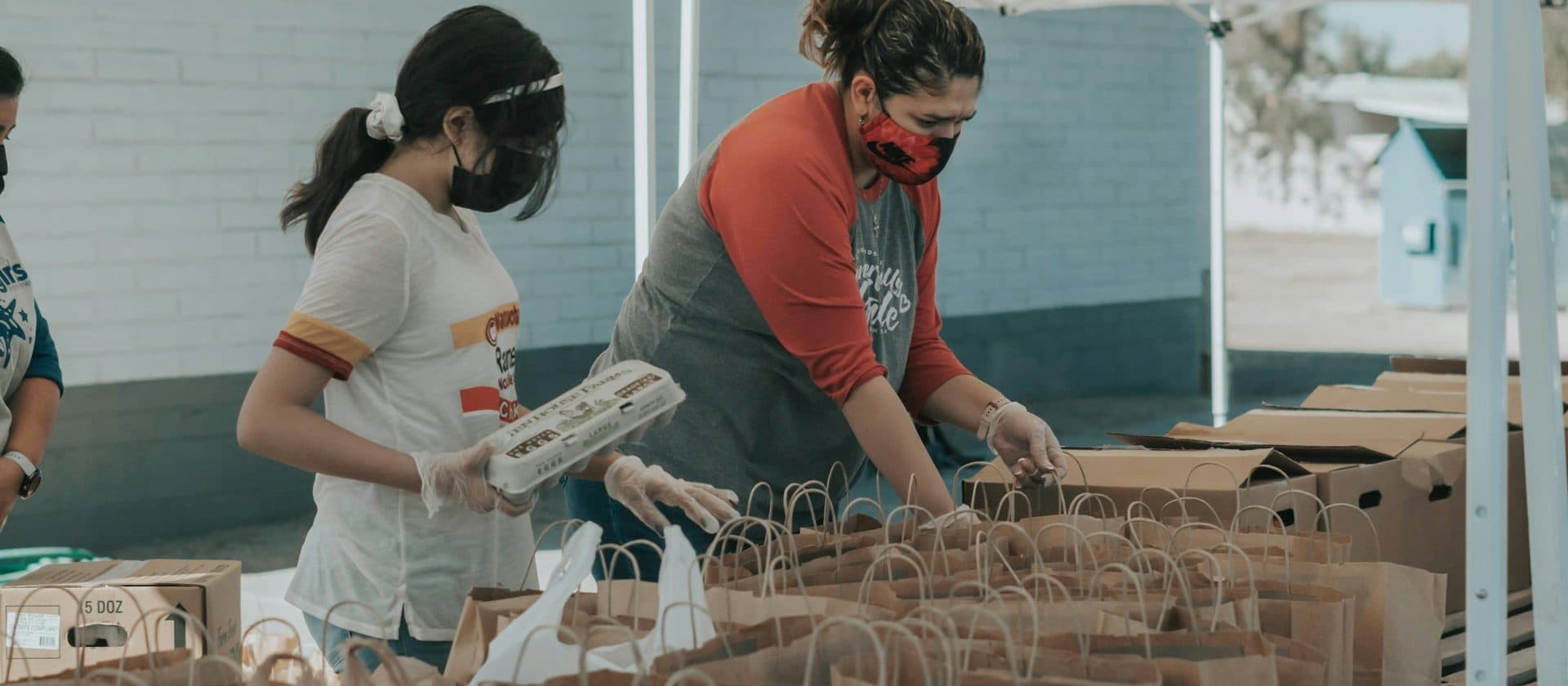 Two volunteers in masks and gloves packing paper bags at a community distribution under a canopy.