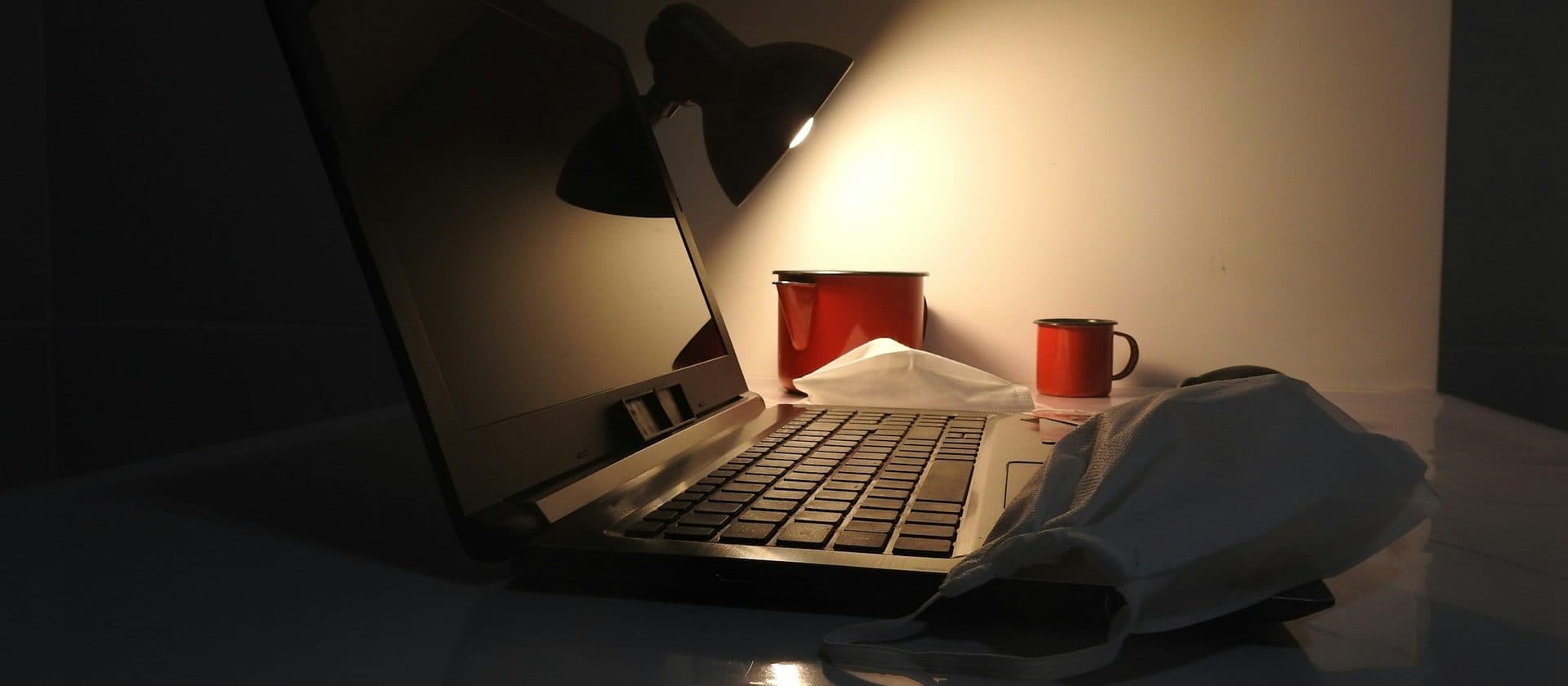 A laptop open under a desk lamp late at night, two enamel mugs beside it.